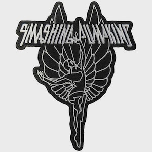 May include: A black and white embroidered patch featuring a stylized winged figure with arms outstretched. The text SMASHING PUMPKINS is embroidered in white across the top section of the patch, which has a distinct black border and white outlines.