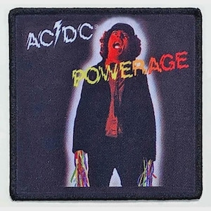 May include: A square fabric patch featuring the AC/DC logo in white and the word POWERAGE in yellow text. The design depicts a person in a dark jacket with a red-tinted face and colourful, string-like elements hanging from the sleeve cuffs.