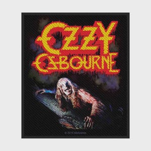 Puede incluir: Un parche de tela rectangular con el texto amarillo Ozzy Osbourne en una fuente que parece gotear. Debajo del texto hay una ilustración gráfica de una criatura de piel pálida y pelo largo con dientes afilados sobre un tronco oscuro y texturizado.