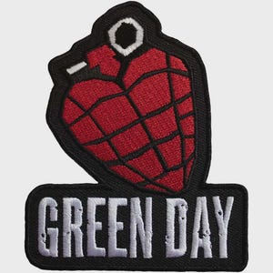 Peut inclure: Un écusson brodé représentant une grenade à main en forme de cœur rouge avec des lignes de grille noires et une goupille blanche. Sous la grenade, une base rectangulaire noire affiche le texte blanc GREEN DAY dans une police sans empattement.
