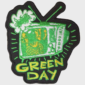 Puede incluir: Un parche bordado que muestra un televisor con pantalla verde y una forma blanca similar a una nube encima. El televisor tiene dos antenas y el texto GREEN DAY está bordado en amarillo debajo del televisor sobre un fondo negro.