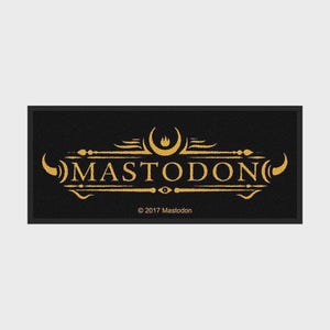 Può includere: Una toppa rettangolare in tessuto nero con il testo MASTODON color oro centrato in un carattere stilizzato. Il design include linee dorate decorative, forme a mezzaluna e una piccola icona a fiamma sopra il testo. Il testo 2017 Mastodon è stampato in basso.