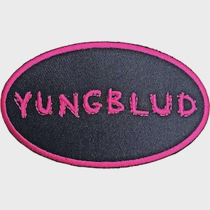 Puede incluir: Un parche bordado de forma ovalada con un fondo de tela negra y un borde cosido en color rosa brillante. El texto YUNGBLUD está bordado en el centro con hilo rosa brillante a juego, utilizando una tipografía estilizada y ligeramente desgastada.