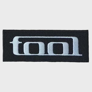 May include: A rectangular embroidered patch featuring a black background with the word TOOL stitched in white thread. The letters are stylized with a bold, blocky font enclosed within a thin white rectangular border that frames the text.