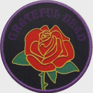 Puede incluir: Un parche bordado circular que presenta una rosa roja central con contornos amarillos y hojas verdes sobre un fondo negro. El texto GRATEFUL DEAD está bordado en morado a lo largo del borde superior curvo, con un borde morado alrededor.