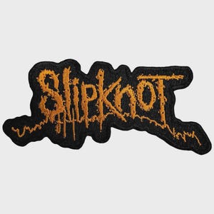 May include: A black embroidered patch featuring the word Slipknot in a jagged, distressed orange font. The lettering has a textured, stitched appearance with uneven edges, set against a solid black background that follows the contour of the text.