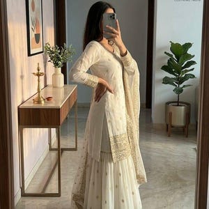 May include: White Indian salwar kameez with gold embroidery on the sleeves, hem, and dupatta. The outfit includes a long-sleeved tunic, wide-legged pants, and a matching scarf. The outfit is worn in a well-lit room with a mirror.