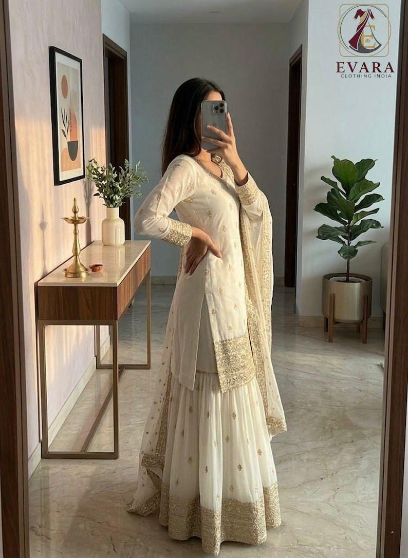 May include: White Indian salwar kameez with gold embroidery on the sleeves, hem, and dupatta. The outfit includes a long-sleeved tunic, wide-legged pants, and a matching scarf. The outfit is worn in a well-lit room with a mirror.