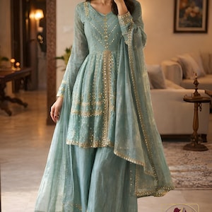May include: A light blue ensemble featuring a tunic top and wide-leg pants, embellished with gold accents. The outfit includes a matching sheer scarf. The clothing is from Evara Clothing.