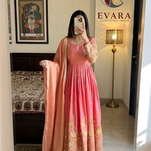 May include: A pink Indian Anarkali suit with gold embroidery on the sleeves and hem. The outfit includes a matching dupatta and pants. The image also shows a framed artwork and a lamp. The brand logo "EVARA CLOTHING INDIA" is visible.