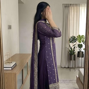 May include: A deep purple salwar kameez ensemble with gold embroidery. The outfit includes a long tunic, matching pants, and a dupatta. The tunic and pants feature intricate gold detailing along the edges and scattered across the fabric. The outfit is from Evara Clothing India.