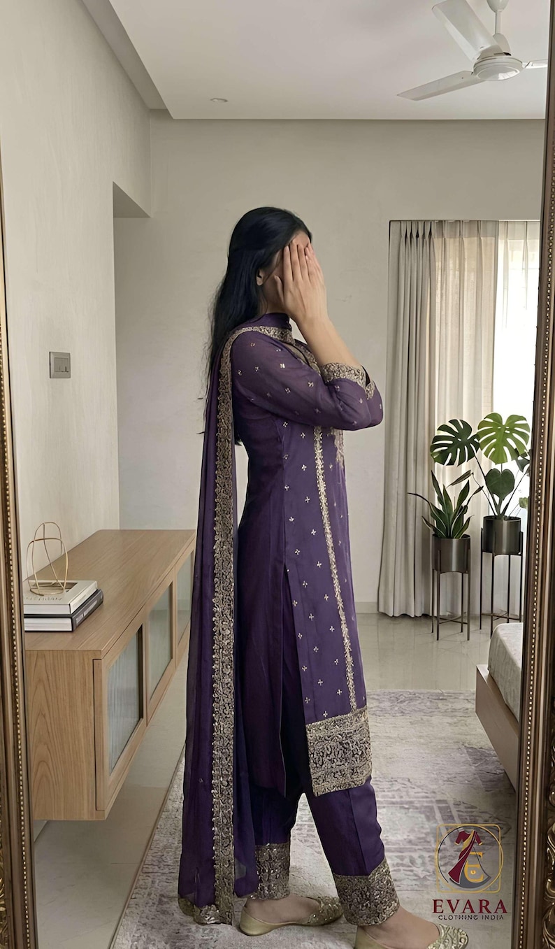May include: A deep purple salwar kameez ensemble with gold embroidery. The outfit includes a long tunic, matching pants, and a dupatta. The tunic and pants feature intricate gold detailing along the edges and scattered across the fabric. The outfit is from Evara Clothing India.