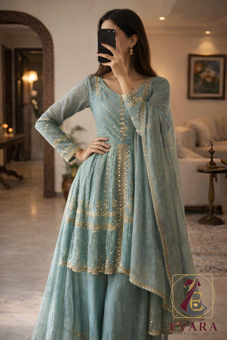 May include: A light blue formal dress with gold embroidery. The dress features a V-neck, long sleeves, and a flowing skirt. The dress is part of a two-piece set and includes a matching scarf. The dress has the text "EVARA CLOTHING NO 4".