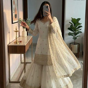 May include: Ivory-colored ethnic wear ensemble with gold embellishments. The outfit includes a long-sleeved tunic, wide-legged pants, and a matching scarf. The trim and details feature gold embroidery. The outfit is suitable for special occasions.