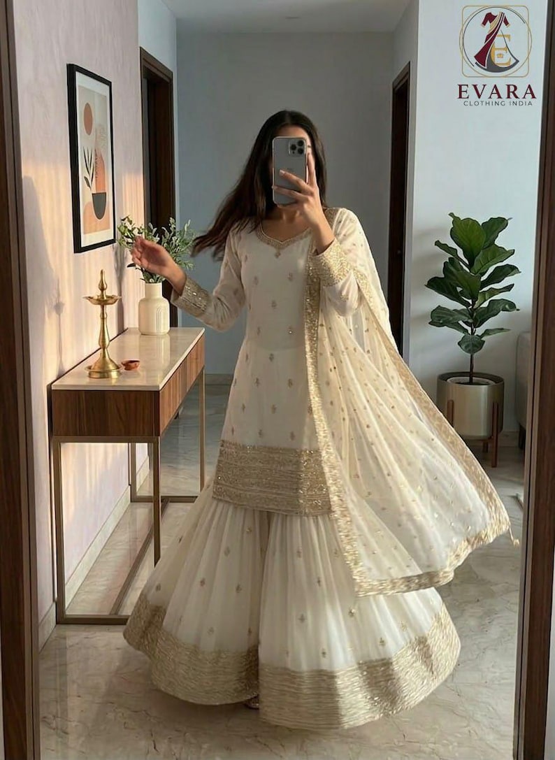 May include: Ivory-colored ethnic wear ensemble with gold embellishments. The outfit includes a long-sleeved tunic, wide-legged pants, and a matching scarf. The trim and details feature gold embroidery. The outfit is suitable for special occasions.