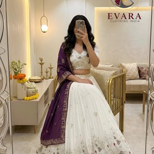 Puede incluir: Conjunto de falda y top lehenga bordados en blanco y dorado con falda plisada. Un chal de color púrpura oscuro con bordados dorados está drapeado sobre los hombros. El atuendo es de EVARA CLOTHING INDIA.