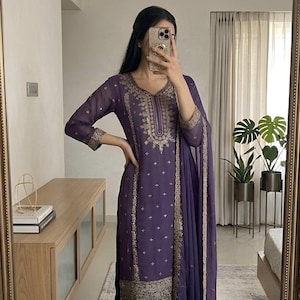 May include: A purple embroidered salwar kameez set with a matching dupatta. The outfit features intricate gold embroidery along the neckline, sleeves, and hem. The image also shows the brand logo "EVARA CLOTHING INDIA".