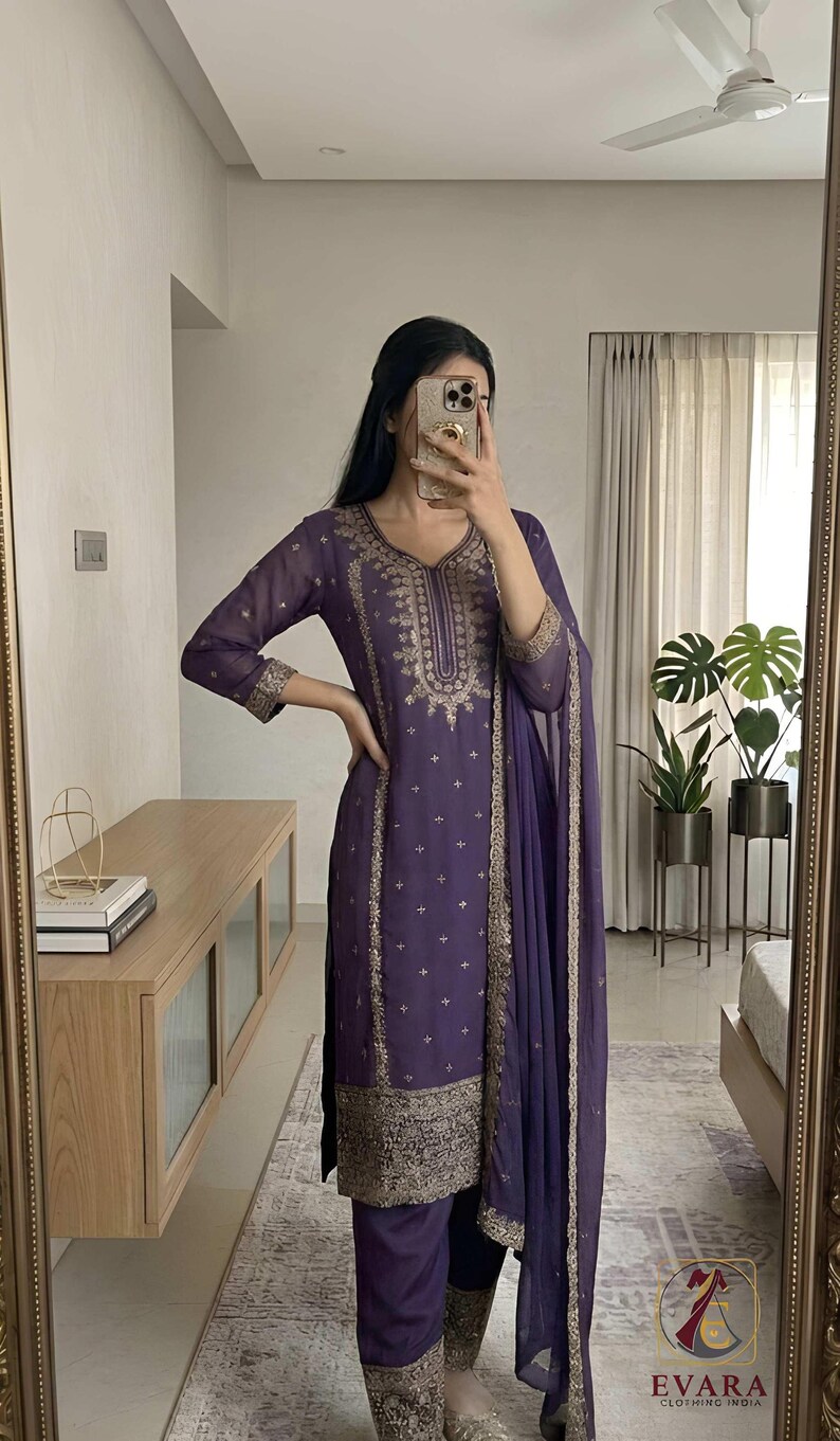 May include: A purple embroidered salwar kameez set with a matching dupatta. The outfit features intricate gold embroidery along the neckline, sleeves, and hem. The image also shows the brand logo "EVARA CLOTHING INDIA".