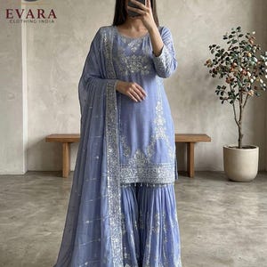 Puede incluir: Un conjunto salwar kameez indio azul claro con intrincados bordados plateados. El atuendo incluye una túnica de manga larga, pantalones de pernera ancha y una bufanda a juego. El logotipo de la marca "EVARA CLOTHING INDIA" es visible.