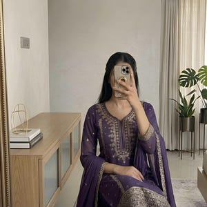 May include: A purple embroidered salwar kameez set. The ensemble features intricate gold embroidery on the neckline, sleeves, and pant hems. The outfit includes a matching dupatta and pants. The clothing is made of a flowing fabric.
