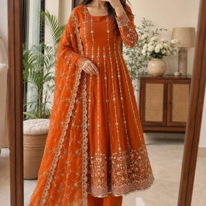 May include: A vibrant orange Anarkali suit featuring a long-sleeved floor-length tunic with vertical silver embroidery and a heavily embellished hem. The set includes matching orange trousers and a sheer orange dupatta with intricate silver floral patterns.