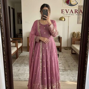 May include: A mauve floor-length anarkali dress featuring a round neckline, long sleeves with intricate embroidery, and a matching dupatta. The garment is adorned with gold-toned floral motifs and a wide, detailed embroidered border at the hem.