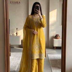 May include: Mustard yellow salwar kameez set with intricate embroidery and embellishments. The set includes a long-sleeved tunic, wide-leg pants, and a sheer dupatta. The outfit is reflected in a large, arched mirror. The brand logo "EVARA CLOTHING INDIA" is visible.