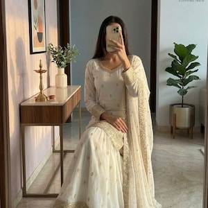 May include: Off-white Indian ethnic wear ensemble. The outfit includes a long-sleeved tunic, wide-legged pants, and a matching scarf, all adorned with gold embroidery. The outfit is worn with matching shoes. The image also includes the logo for Evara Clothing India.