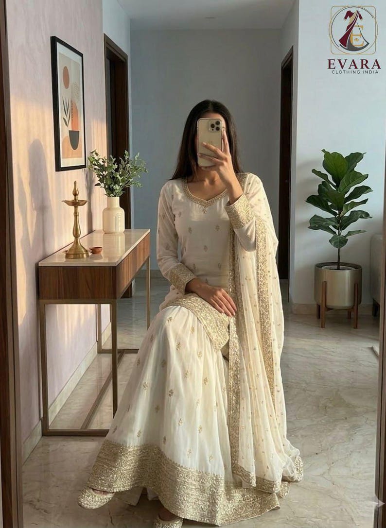 May include: Off-white Indian ethnic wear ensemble. The outfit includes a long-sleeved tunic, wide-legged pants, and a matching scarf, all adorned with gold embroidery. The outfit is worn with matching shoes. The image also includes the logo for Evara Clothing India.