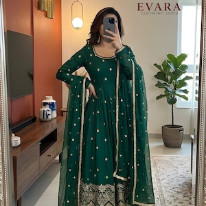 May include: Emerald green Indian salwar kameez with gold embroidery and embellishments. The long-sleeved dress features a round neckline and a flowing silhouette, paired with matching pants and a sheer dupatta. The outfit is from Evara Clothing India.