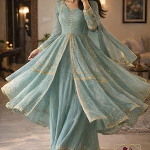 May include: A flowing, light blue dress with gold embellishments. The dress features long sleeves, a V-neck, and a split front, with gold trim outlining the edges. The dress is floor-length and has a matching sheer overlay. The image also shows the logo for "EVARA CLOTHING ND 4."