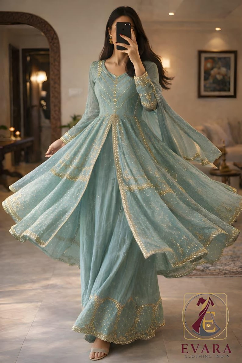 May include: A flowing, light blue dress with gold embellishments. The dress features long sleeves, a V-neck, and a split front, with gold trim outlining the edges. The dress is floor-length and has a matching sheer overlay. The image also shows the logo for "EVARA CLOTHING ND 4."