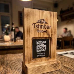 Può includere: Un cartello rettangolare in legno con finitura bruciata che presenta il testo inciso Tsimbar Bistrot e un logo. Un inserto quadrato nero al centro mostra un codice QR bianco sopra il testo Scansiona per il Menu su una base in legno.