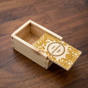 Könnte beinhalten: Eine rechteckige, helle Holzschachtel mit einem Schiebedeckel. Der Deckel ist mit einem goldfarbenen, verzierten Blumenmuster und einem zentralen kreisförmigen Emblem versehen, das den Text "your personalized logo" in Dunkelbraun enthält.