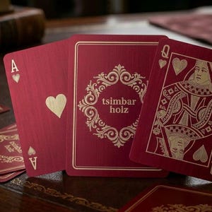 Puede incluir: Un juego de cartas con un acabado de metal cepillado en rojo intenso y grabados dorados. Las cartas muestran el texto tsimbar holz dentro de un marco circular ornamentado, junto con las caras del As de corazones y la Reina de corazones.