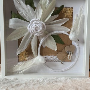 May include: A white framed shadow box art piece featuring a large paper flower with white petals and a crocheted center. The artwork includes green paper leaves, white feathers, gold accents, and a key charm.