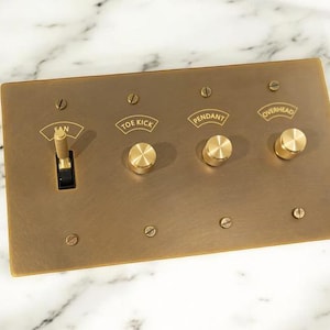 Op de afbeelding: Een rechthoekige messing lichtschakelaarplaat met een tuimelschakelaar en drie draaiknoppen. De plaat is gemarkeerd met "FAN", "TOE KICK", "PENDANT" en "OVERHEAD". De plaat is met schroeven bevestigd.