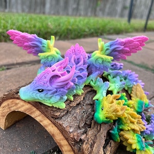 May include: A colorful, articulated dragon figurine in shades of purple, pink, green, and yellow. The dragon is posed on a piece of wood, with its wings spread. The dragon's head is detailed with small black eyes.
