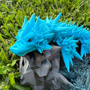 May include: A detailed, 3D-printed dragon sculpture. The dragon features a light blue, spiky head and spine, with a dark gray, geometric body. The sculpture is set against a backdrop of green grass.