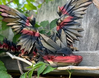 May include: A red, black, and gold dragon sculpture with wings spread wide. The dragon is perched on a wooden branch with green leaves.