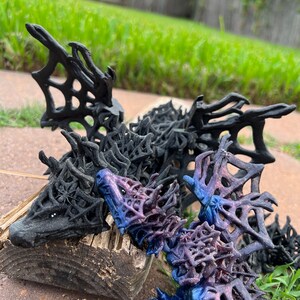 May include: Two 3D printed dragon figurines, one black and one purple with a glittery finish. The dragons are posed with their wings spread out and their heads facing forward. The dragons are made of a lattice-like material.
