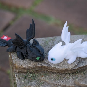May include: Two dragon figurines, one black and one white, rest on a stone surface. The black dragon has green eyes and a red and white tail fin. The white dragon has blue eyes. Both have detailed wings and are in a relaxed pose.