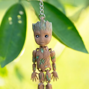 May include: A brown and green Groot keychain, featuring a detailed 3D-printed design. The keychain is suspended from a silver chain and ring, with a blurred green background.
