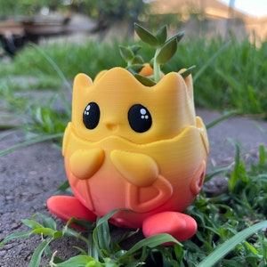 May include: A yellow and orange gradient ceramic planter shaped like a cartoon character with large eyes and a small mouth. The planter has two red feet and is holding a small green succulent plant.