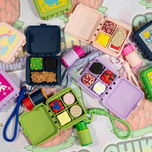 May include: A collection of miniature square lunchbox containers in various colors including pink, blue, purple, and green. Each container features a hinged lid with graphic designs like unicorns, rainbows, and rockets, and contains small food replicas.