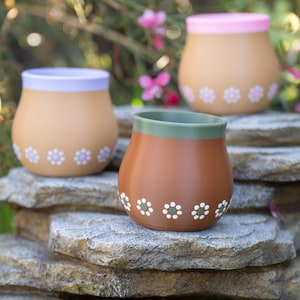 May include: Three ceramic pots with floral designs. The pots are in shades of brown, tan, and a light purple, with white and green floral patterns. The rims are painted in green, pink, and light purple. The pots are displayed on a stone surface.