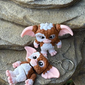 May include: Two Gremlins figurines, one standing and one lying down, with brown, white, and pink details. The standing figurine has a keychain attachment. The figures have large ears and expressive faces. The background is a textured, light-colored stone.