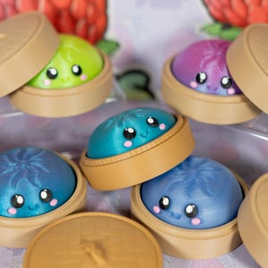May include: A collection of colorful, smiling dumpling toys in bamboo steamers. The dumplings are various colors, including green, blue, and purple, with cute, cartoonish faces. The steamers are tan with a textured lid.
