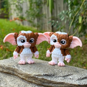 May include: Two small, detailed figurines of a Gremlin character. The figures have brown fur, white bellies, pink ears and feet, and large black eyes. They are standing on a gray rock, with a blurred green background of foliage.
