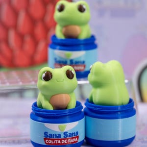 May include: Three green frog figurines in blue containers. Each frog has large brown eyes and a tan belly. The containers have the text "Sana Sana Colita de Rana" in white and red. The background is blurred.